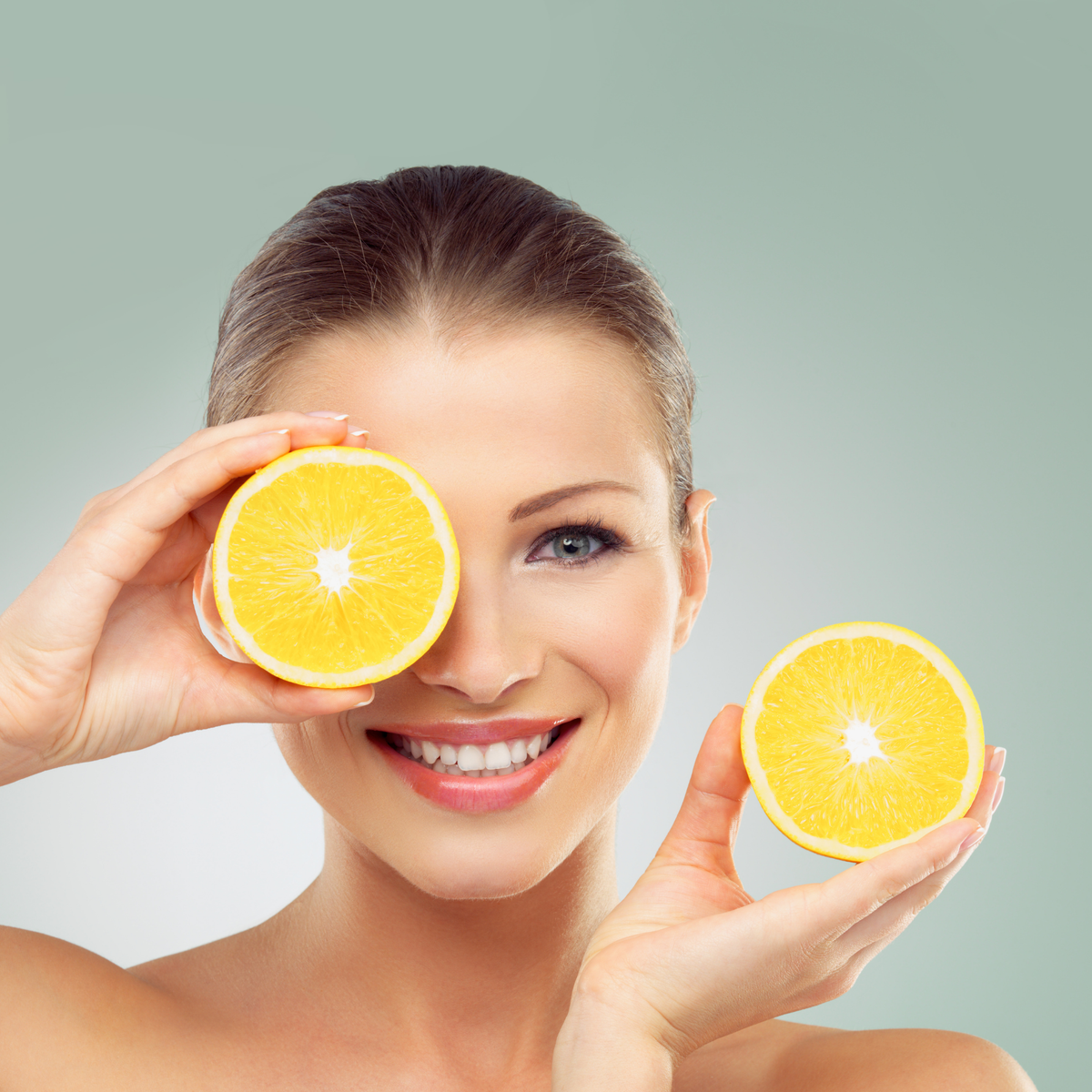 Young woman smiling while holding two orange halves. One in front o fher face, the other on the right hand side of the image. 